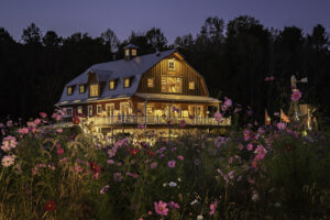 Twilight exterior of a barn style home glowing behind a wildflower meadow.